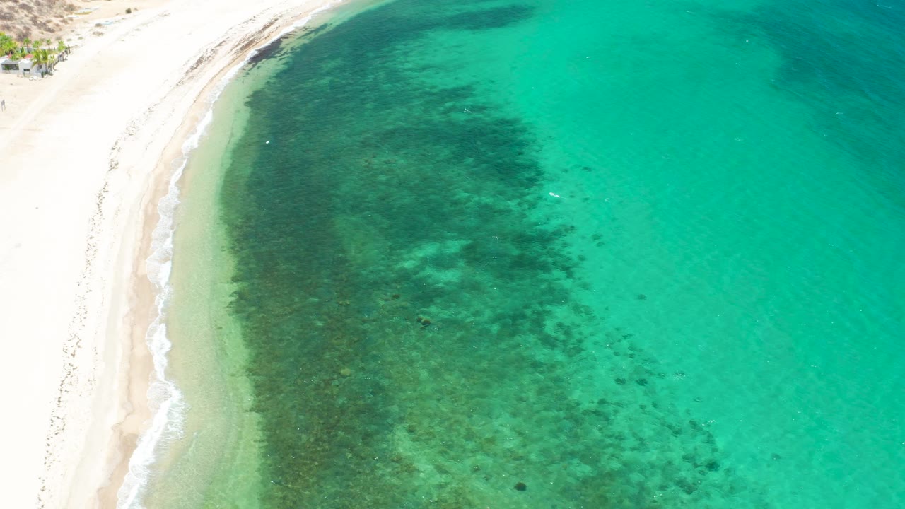 Aerial frame from El Sargento La Ventana, Mexico, showing a slightly higher section of shoreline, pale sand, transparent turquoise shallows, mottled dark seabed texture, and no visible people.