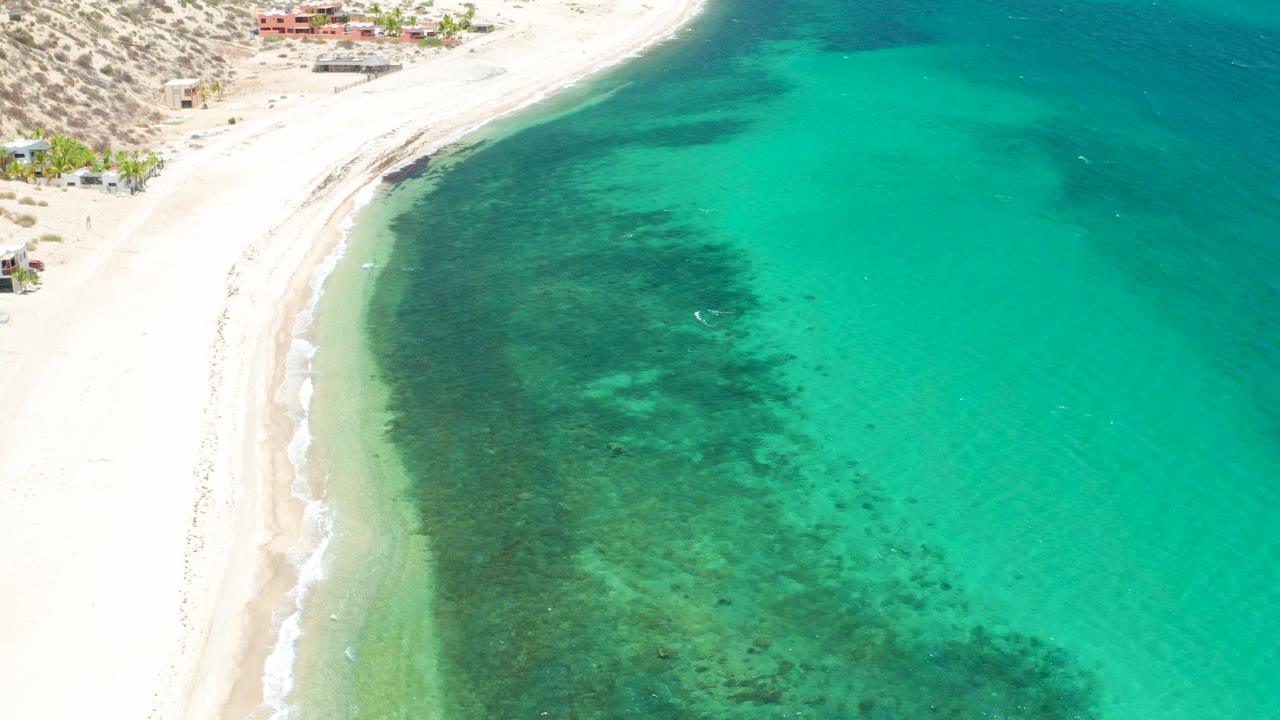 Aerial view over El Sargento La Ventana, Mexico, with a pale beach on the left, turquoise water on the right, darker underwater patches near shore, hillside buildings above the sand, and no visible people.