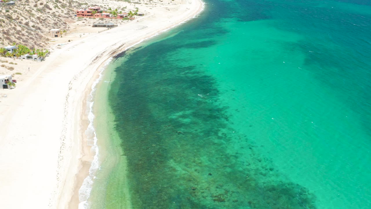 Aerial frame over El Sargento La Ventana, Mexico, showing the beach bend near the top left, wide turquoise water across the frame, a dark underwater band close to shore, and no visible people.