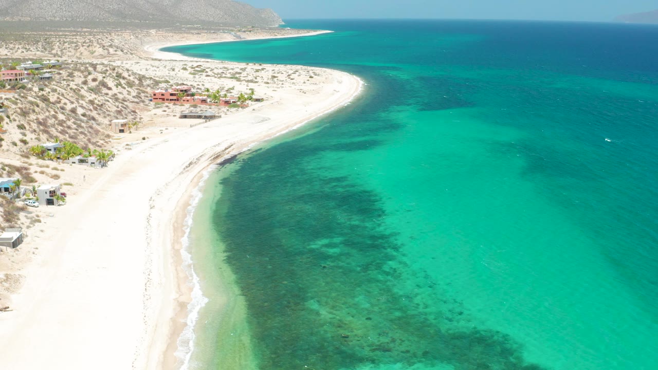 Aerial view in El Sargento La Ventana where turquoise water extends across the frame, a white sand beach curves below desert hillside terrain, and no visible people appear.