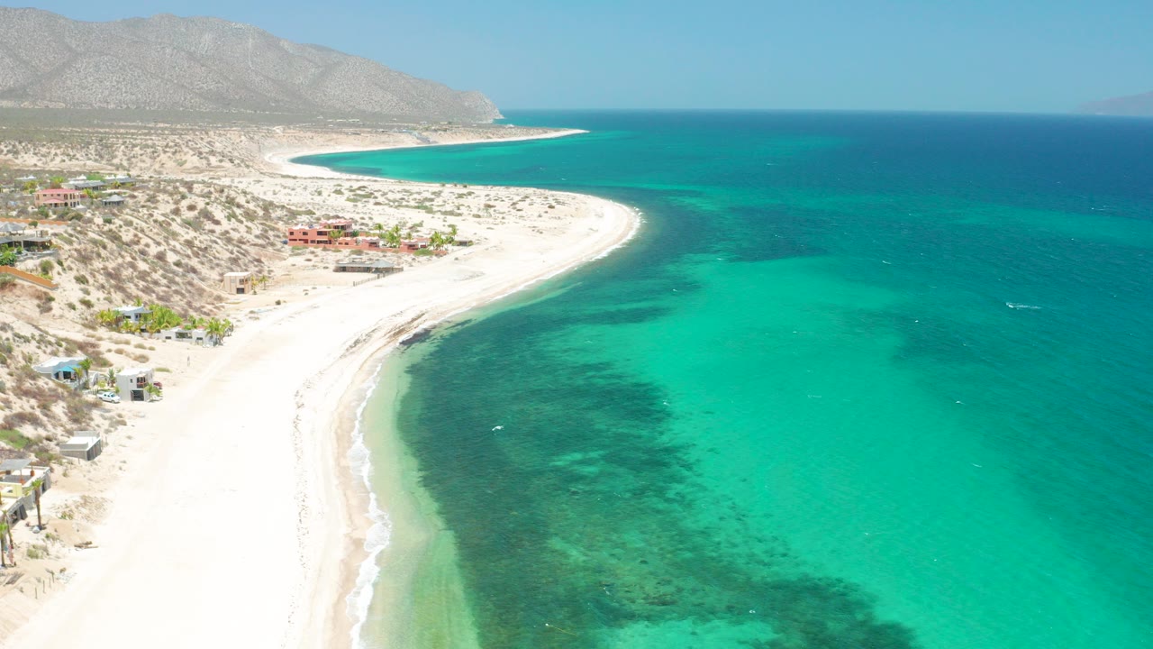 High aerial frame of El Sargento La Ventana, Mexico, showing a curved white sand shoreline, turquoise shallows, darker underwater patches, desert shrubs, small shore buildings, mountains, and no visible people.
