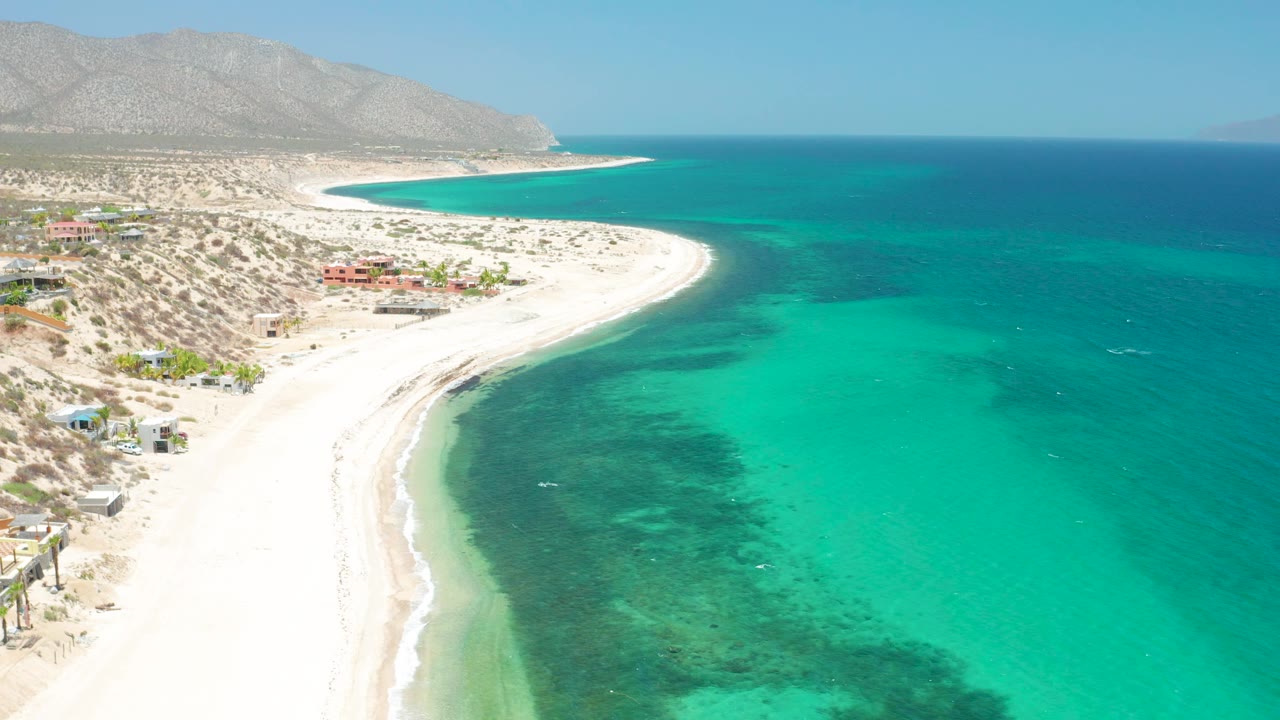 Aerial frame over El Sargento La Ventana, Mexico, with a bright sand spit curving into turquoise water, darker reef or sea grass areas below the surface, sparse shoreline buildings, mountains, and no visible people.