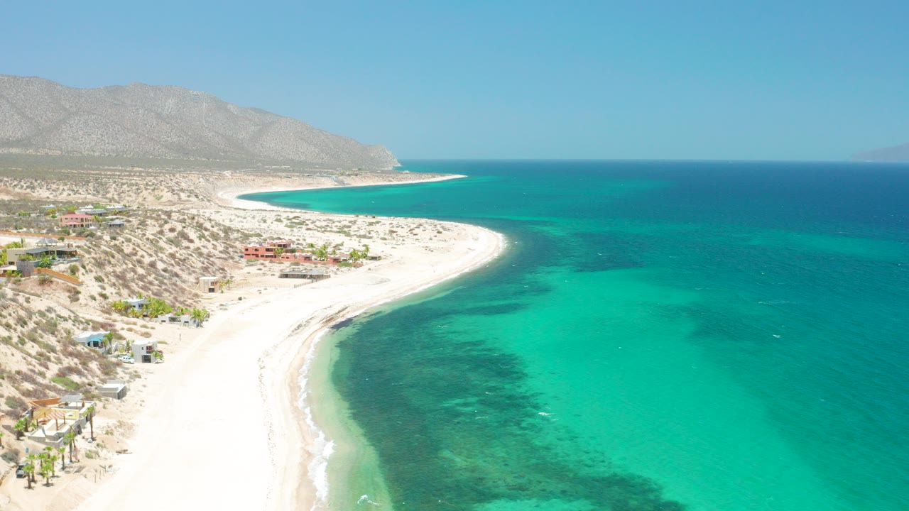 Opening aerial frame over El Sargento La Ventana, Mexico, showing a bright white beach, turquoise shallows, deeper blue water, distant mountains, scattered shoreline buildings, and no visible people.