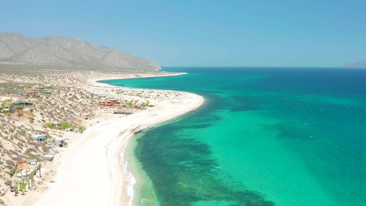 Aerial view over El Sargento La Ventana, Mexico, where more open turquoise water fills the right side, the white beach arcs along the left, mountains sit under clear sky, and no visible people appear.
