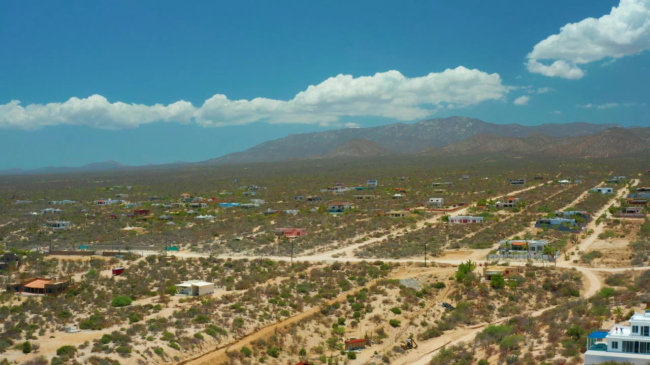 Aerial view across El Sargento La Ventana, Mexico, with dry lots, small homes, winding dirt tracks, hazy mountains, bright blue sky, and no visible people.