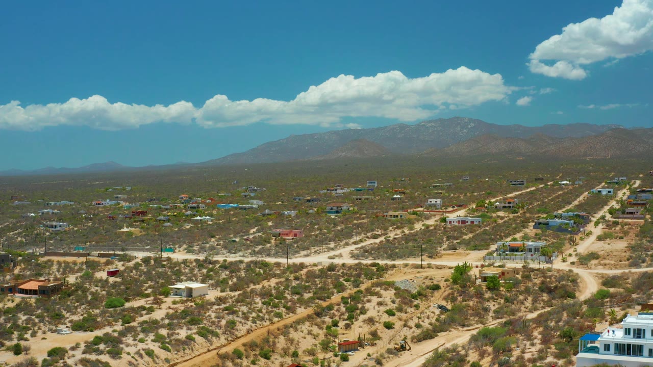 Drone frame over El Sargento La Ventana, Mexico, showing a diagonal dirt road through sparse houses, scrubland, mountain backdrop, bright clouds, and no visible people.