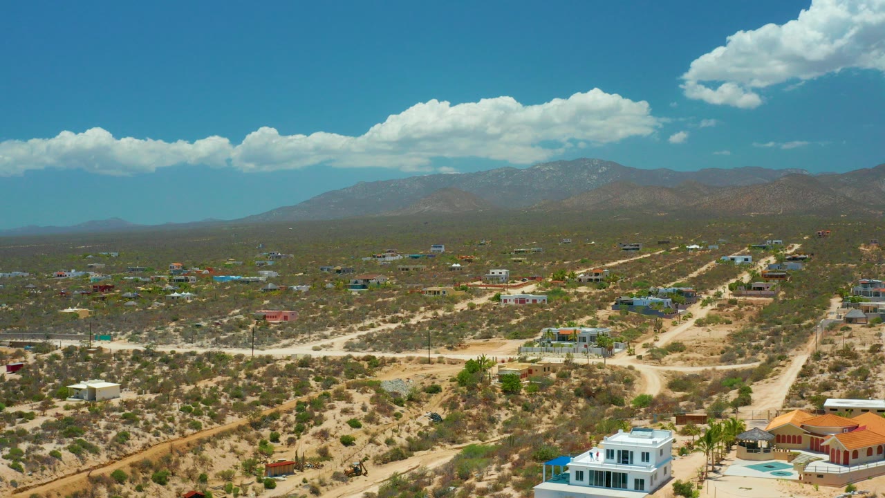 Opening aerial frame over El Sargento La Ventana, Mexico, showing scattered desert houses, pale dirt roads, distant mountains, bright clouds, and no visible people.