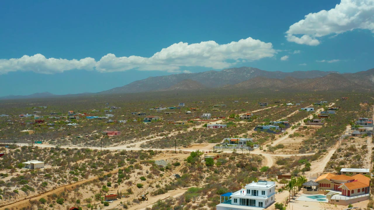 Aerial frame over El Sargento La Ventana, Mexico, looking past foreground homes and open desert lots toward hazy brown mountains, white clouds, and no visible people.