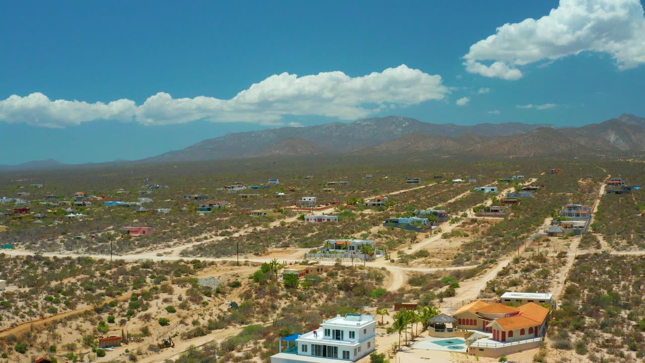 Opening aerial frame over El Sargento La Ventana, Mexico, with large homes on dry lots, pale dirt roads, a mountain horizon, bright clouds, and no visible people.