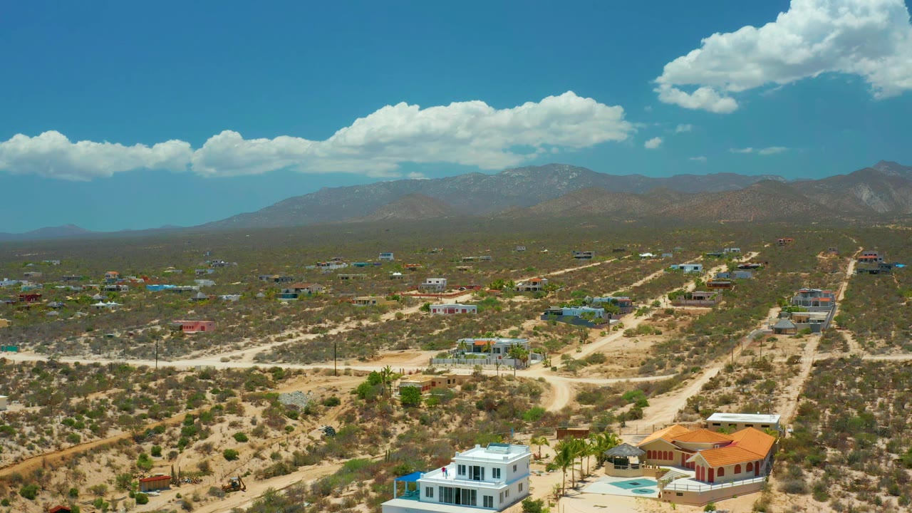 Early frame over El Sargento La Ventana, Mexico, showing scattered houses, intersecting pale roads, dry vegetation, cloud shadows, mountains, and no visible people.