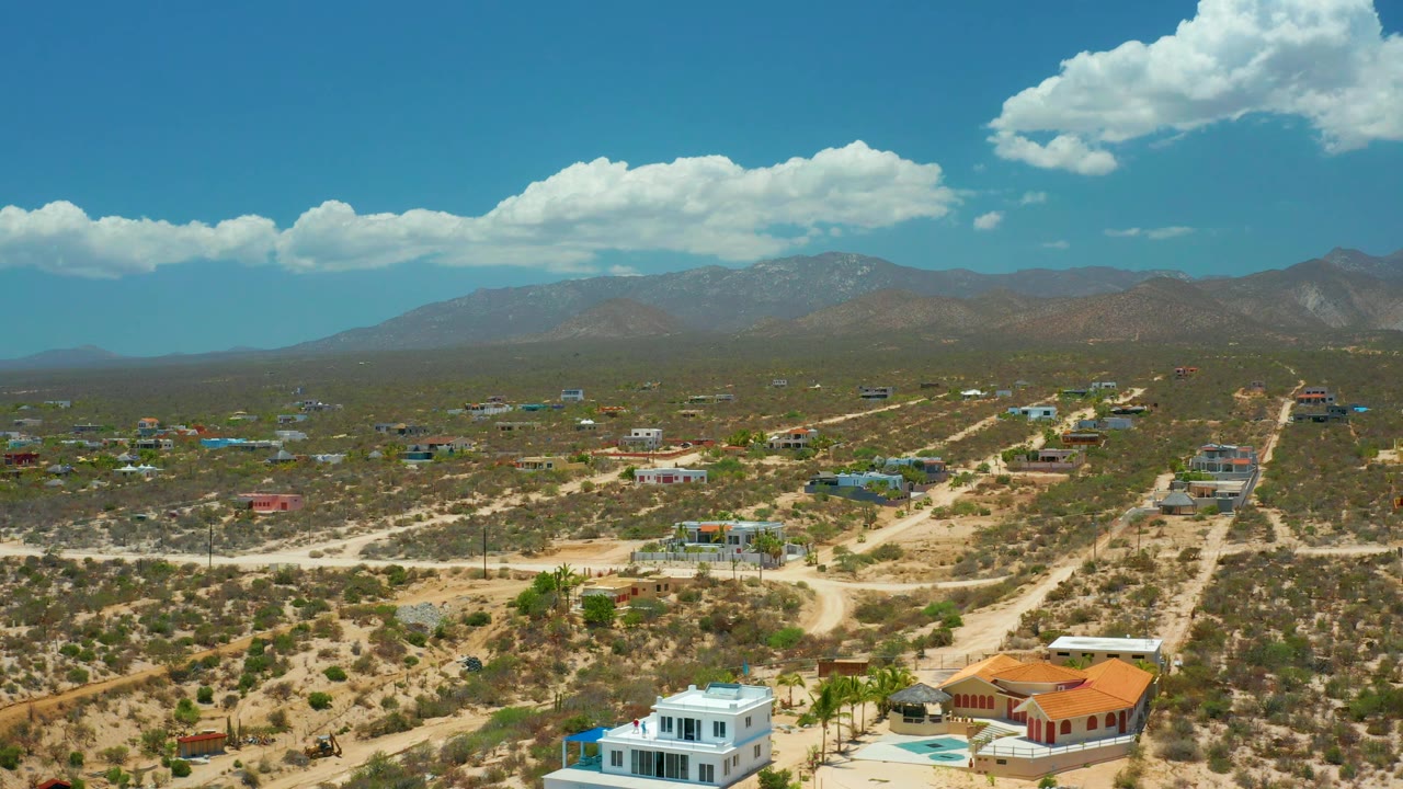 Aerial frame in El Sargento La Ventana, Mexico, with colorful rooflines among dry lots, a white foreground home, winding dirt roads, mountain backdrop, and no visible people.