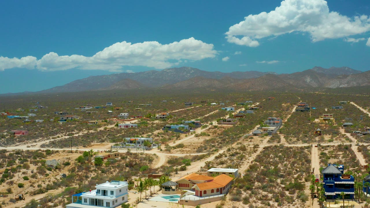 Opening aerial frame over El Sargento La Ventana, Mexico, with dirt roads, low homes, scrubland, distant mountains, bright clouds, and no visible people.