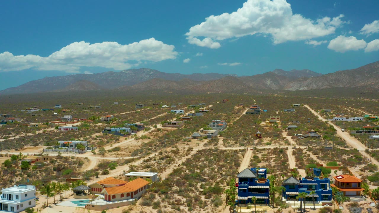 Opening aerial frame over El Sargento La Ventana with large homes at the bottom edge, sandy roads crossing desert brush, distant mountains, bright clouds, and no visible people.