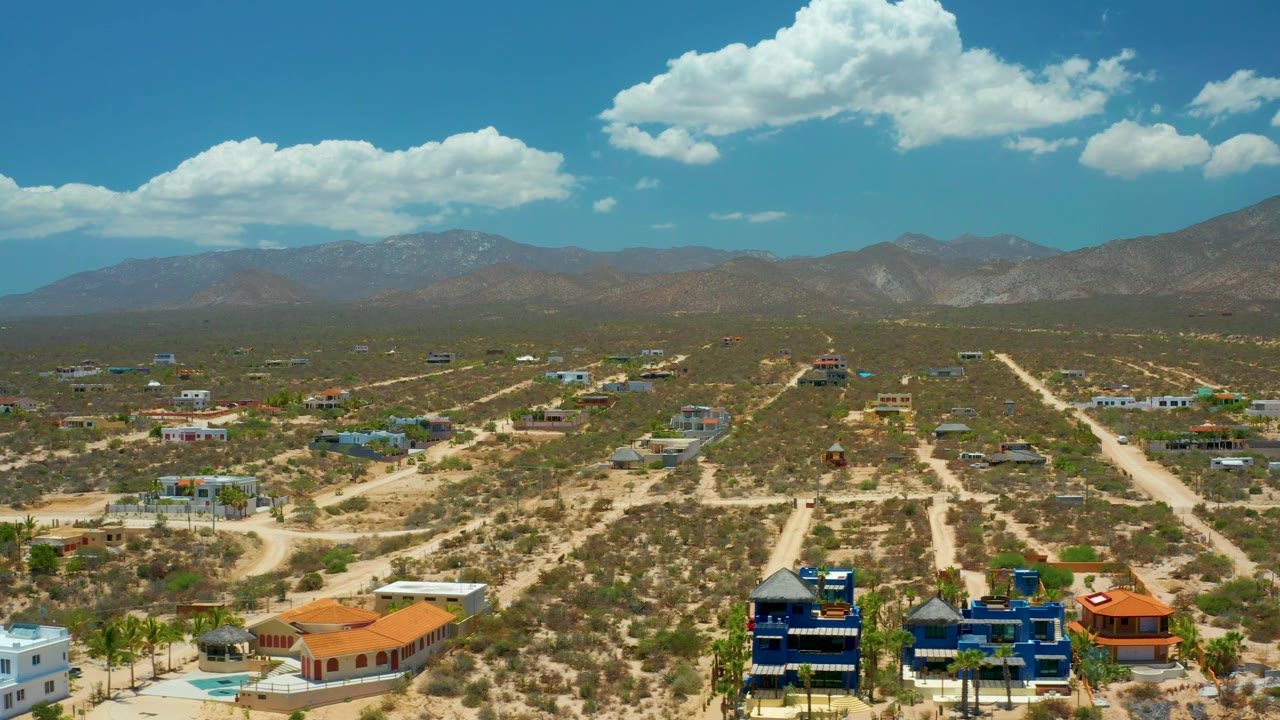 El Sargento La Ventana aerial frame with a blue multi-level home near the lower edge, sandy roads radiating through undeveloped lots, mountains beyond, and no visible people.