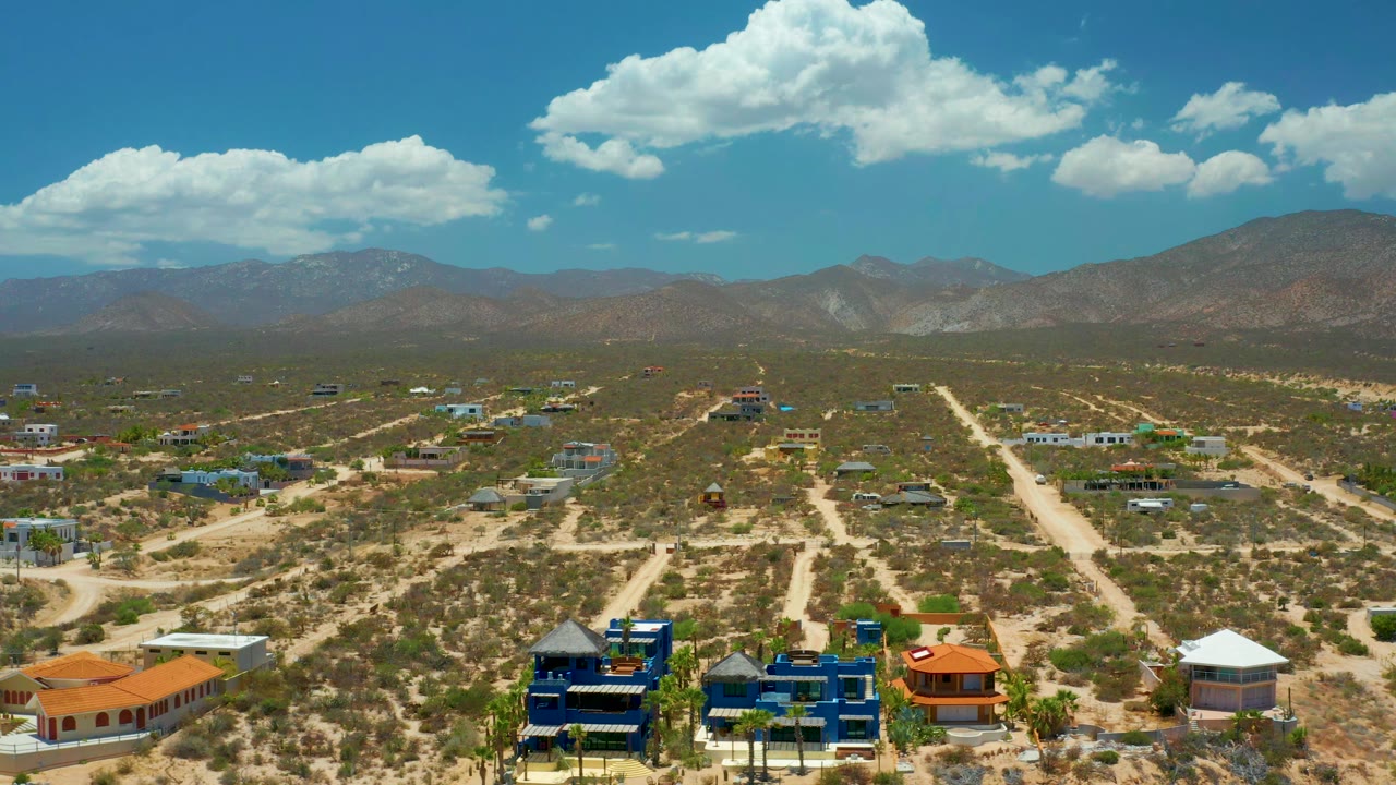 Second aerial frame over El Sargento La Ventana, Mexico, with the dirt-road grid and colorful low buildings in the foreground, mountains beyond, and no visible people.
