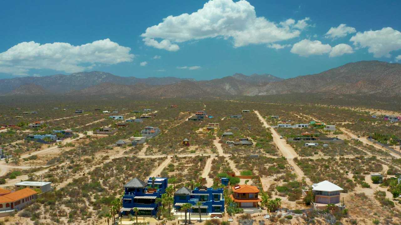 Wide aerial landscape with village rooftops, branching dirt roads, mountains, and clouds.