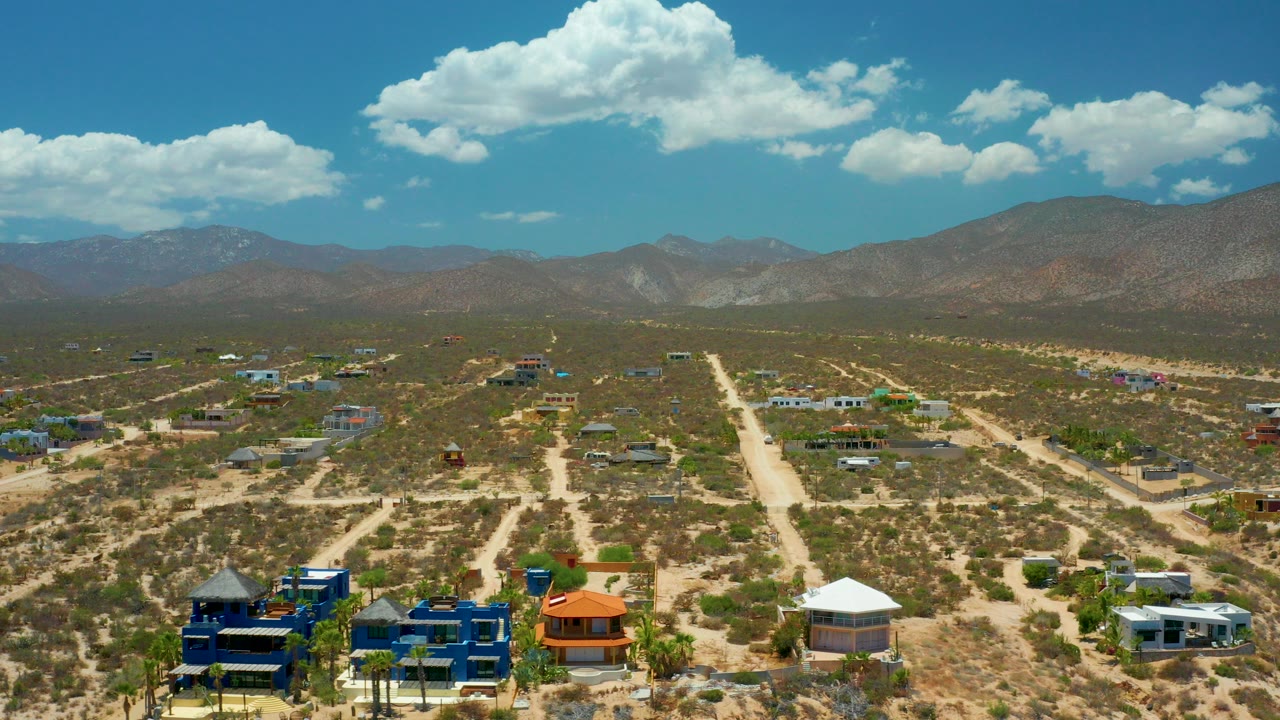 Opening aerial frame over El Sargento La Ventana, Mexico, with scattered homes along dirt roads, scrub-covered terrain, distant mountains, white clouds, and no visible people.
