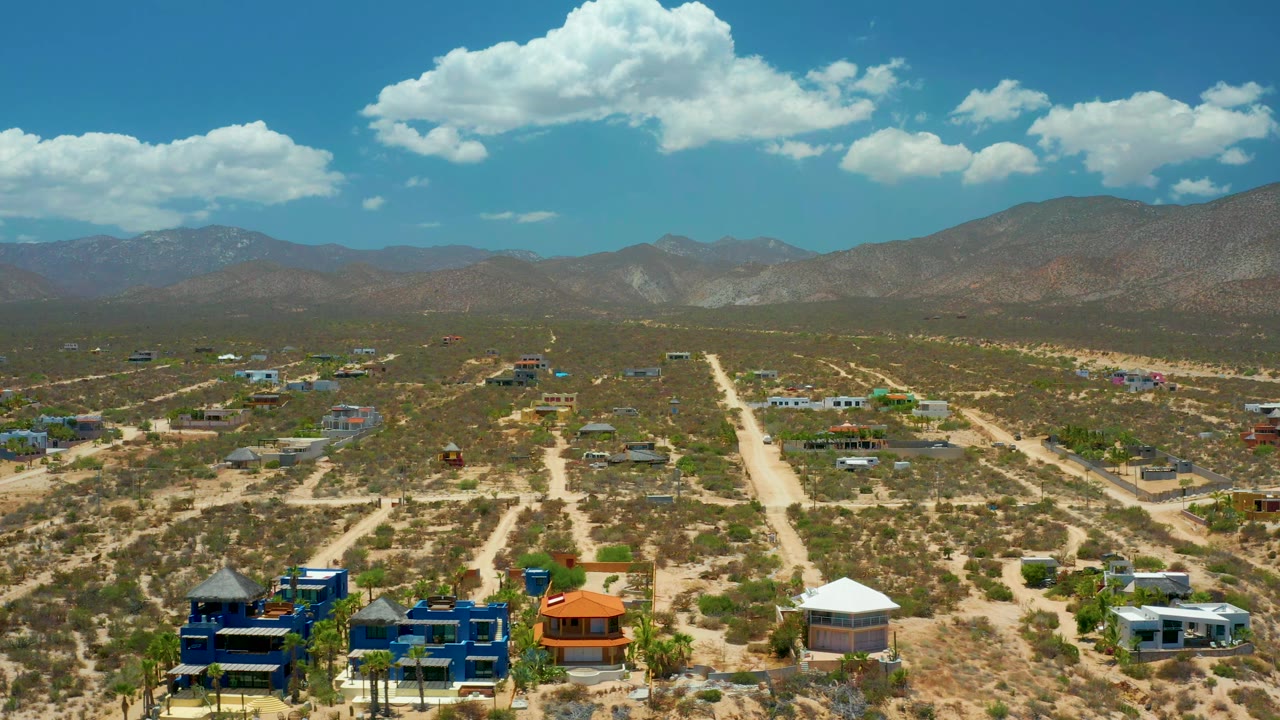 Second frame holds a wide drone view of El Sargento La Ventana with a straight pale dirt road through dry terrain, low buildings across the midground, mountains behind, and no visible people.