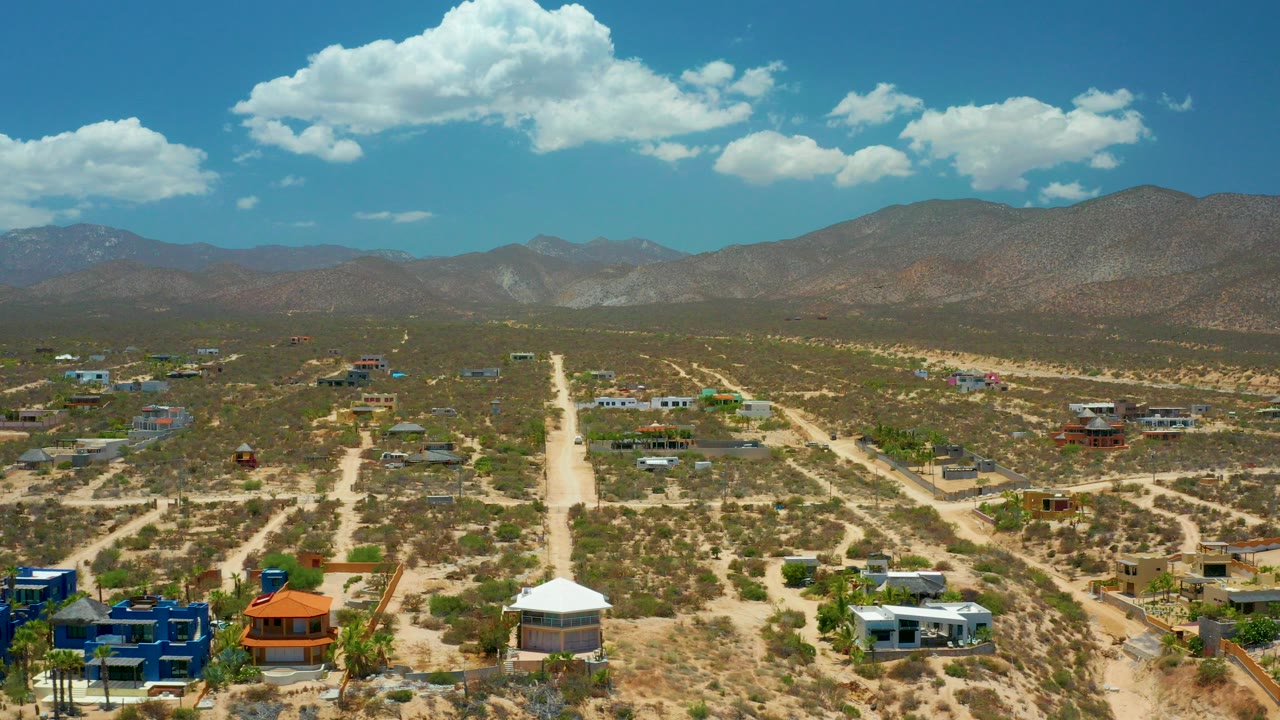 High aerial view over El Sargento La Ventana with a pale unpaved road running toward distant mountains, scattered homes below, bright clouds overhead, and no visible people.