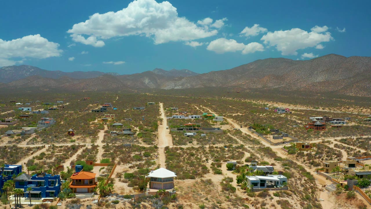 Drone-height view of El Sargento La Ventana showing the central dirt road slightly farther through the frame, low homes across dry lots, brown mountains, and no visible people.
