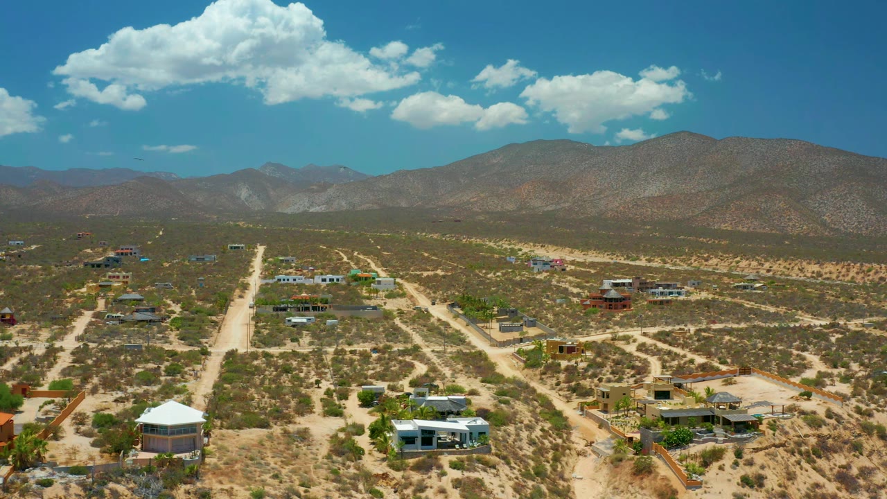 Aerial frame in El Sargento La Ventana, Mexico, with a central sandy road, low buildings spread through brush, mountains on the horizon, and no visible people.