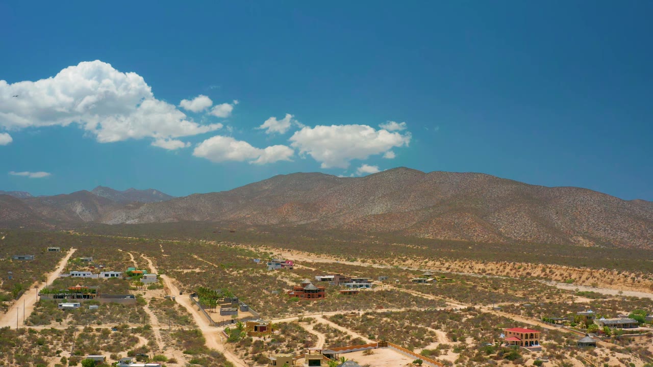 Aerial frame in El Sargento La Ventana showing more of the branching dirt road grid, scattered buildings, scrubland, mountain ridge, bright clouds, and no visible people.