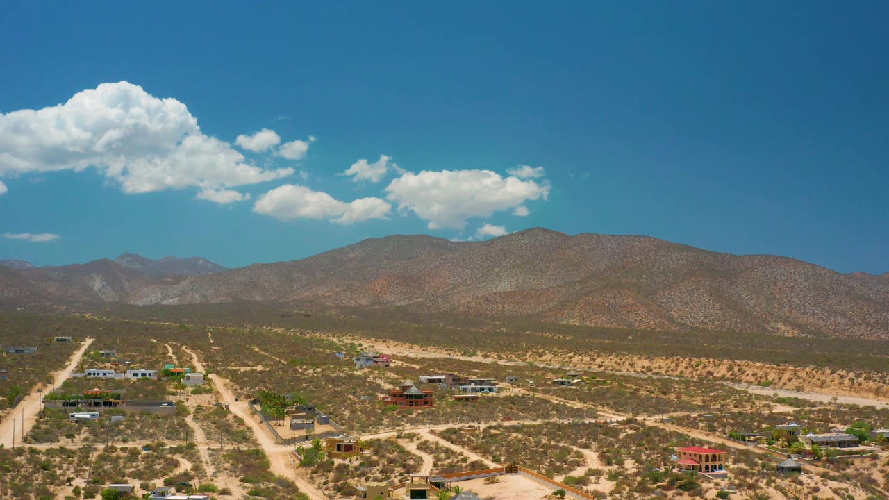 El Sargento La Ventana aerial view with pale roads crossing desert flats, a few homes near the lower frame, brown mountains on the horizon, large clouds, and no visible people.