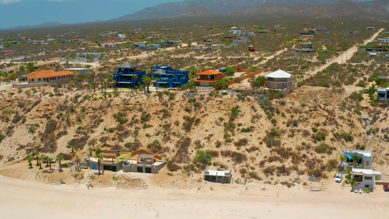 Aerial view in El Sargento La Ventana, Mexico, with pale sand along the bottom, a scrubby bluff with modern homes, dirt roads, distant mountains, and no visible people.