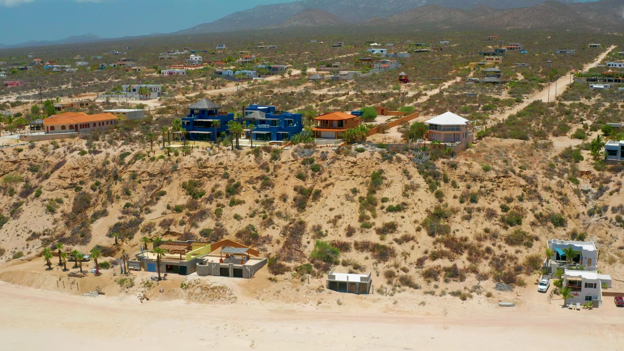 Aerial view over El Sargento La Ventana with a wide strip of pale beach, eroded bluff, scattered homes, sandy roads leading inland, hazy mountains, and no visible people.