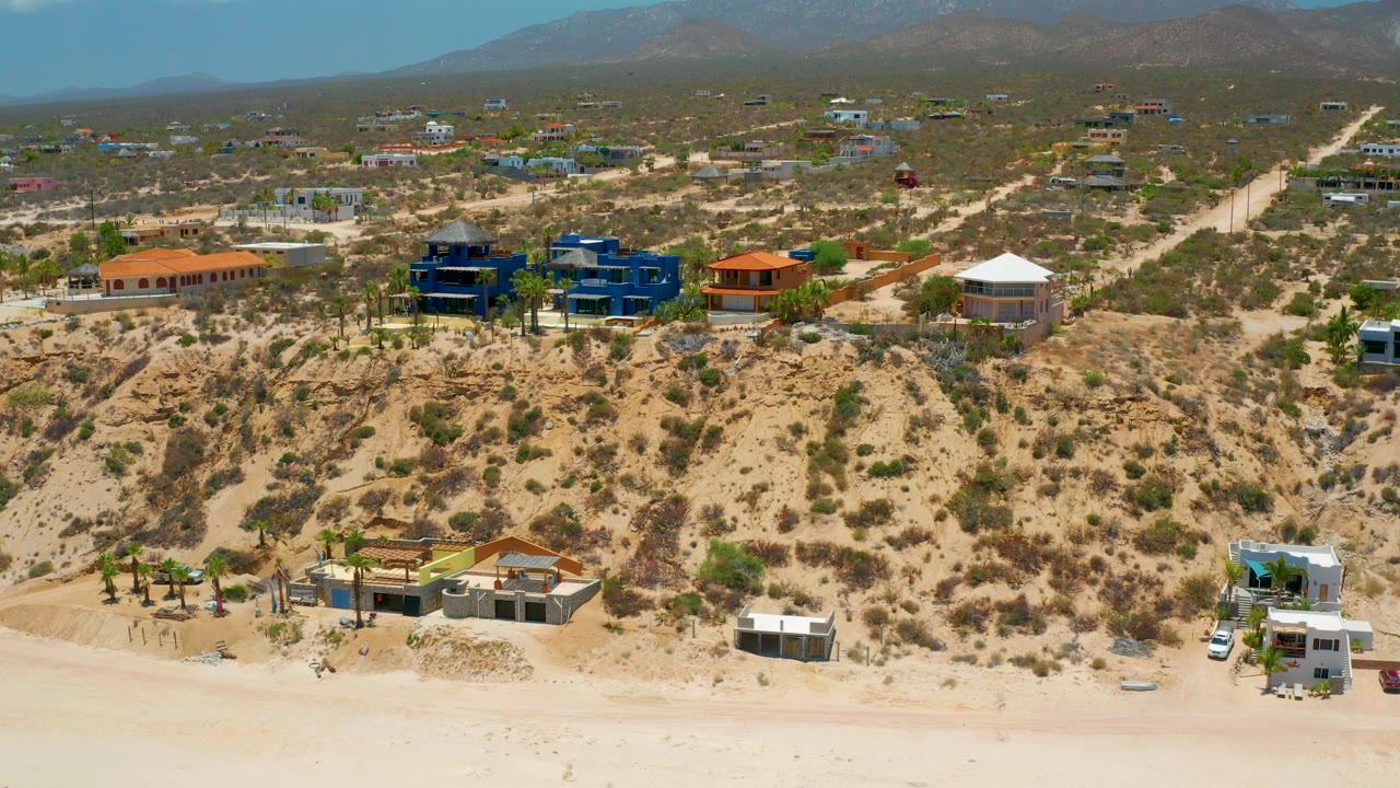 Beach below desert bluff with homes, palms, dirt roads, and mountain horizon.