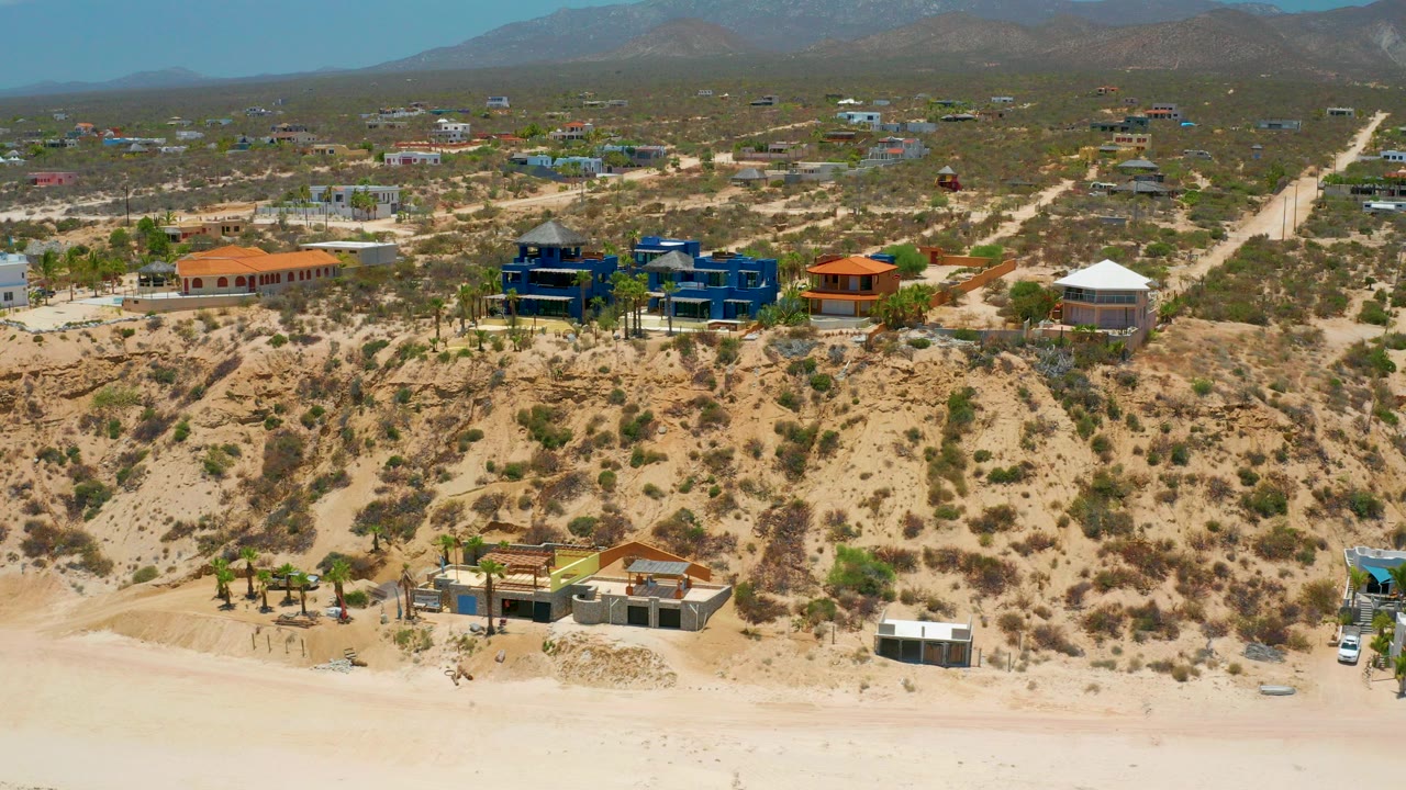 Wide aerial frame over El Sargento La Ventana showing pale beach, scrubby bluff, scattered houses, dirt roads, blue sky, distant mountains, and no visible people.