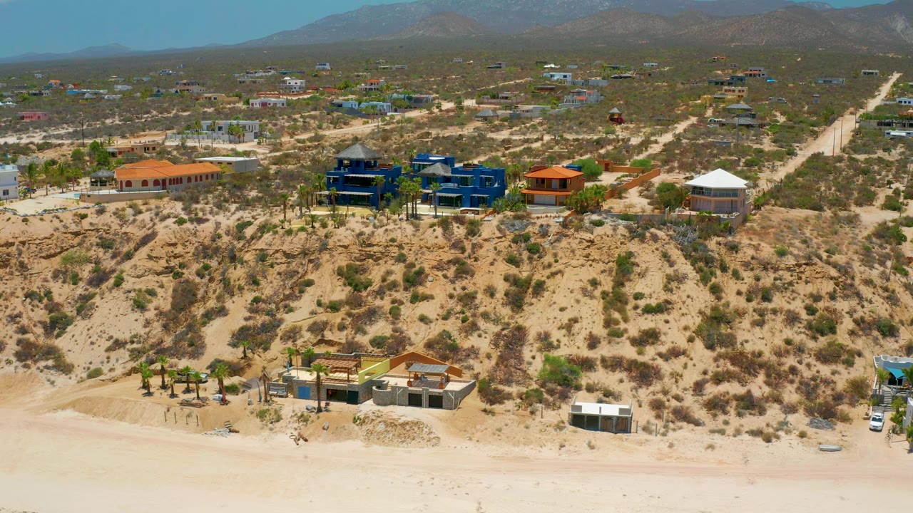 Wide drone view of coastal houses above a sandy beach with mountains behind.