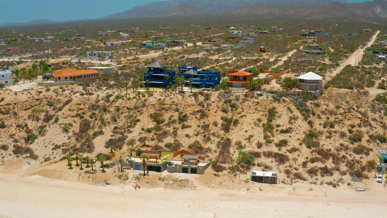 Wide 4K aerial still of El Sargento La Ventana showing homes along a scrubby coastal rise, pale sand in front, open roads behind, and no visible people.