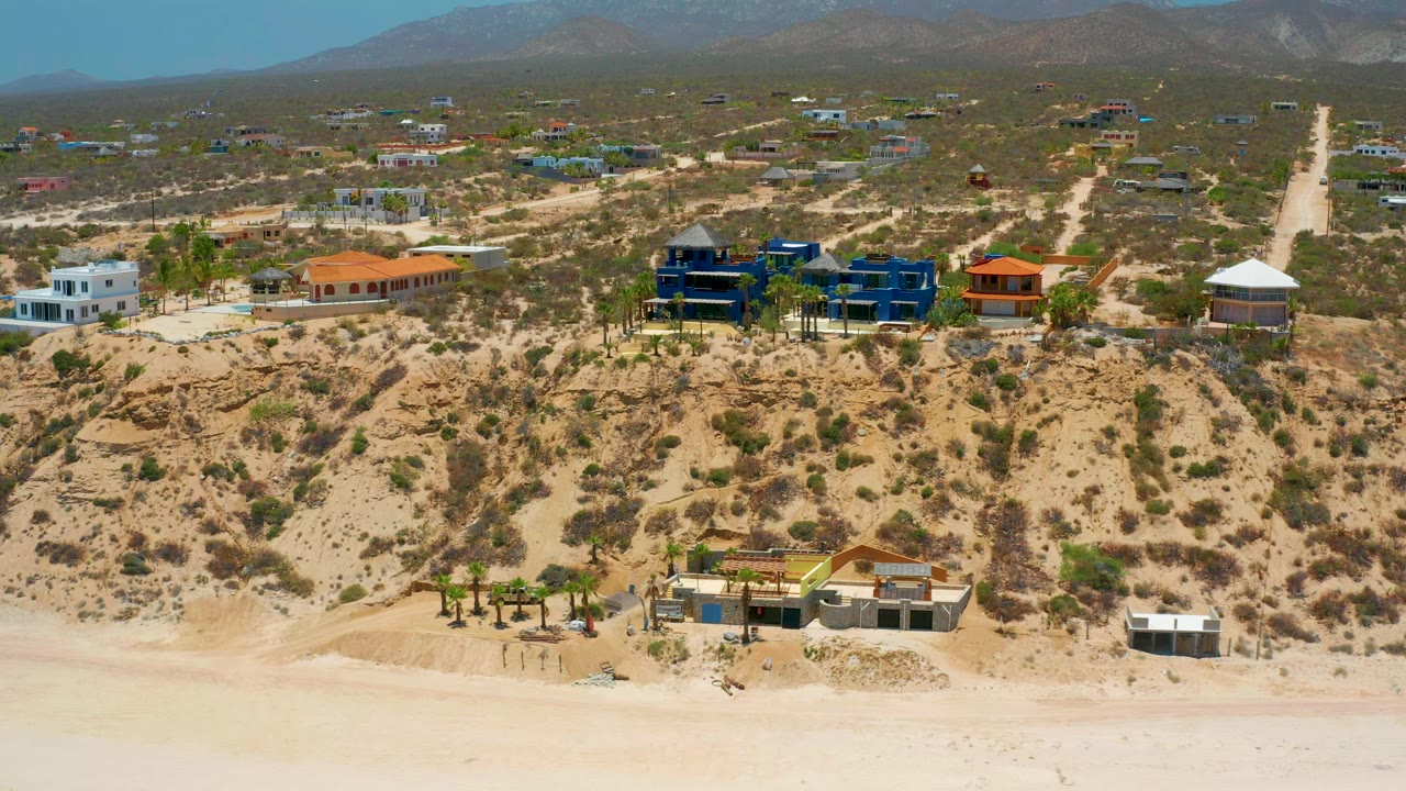 Wide drone view of beach dunes, colorful homes, dirt roads, and mountains.