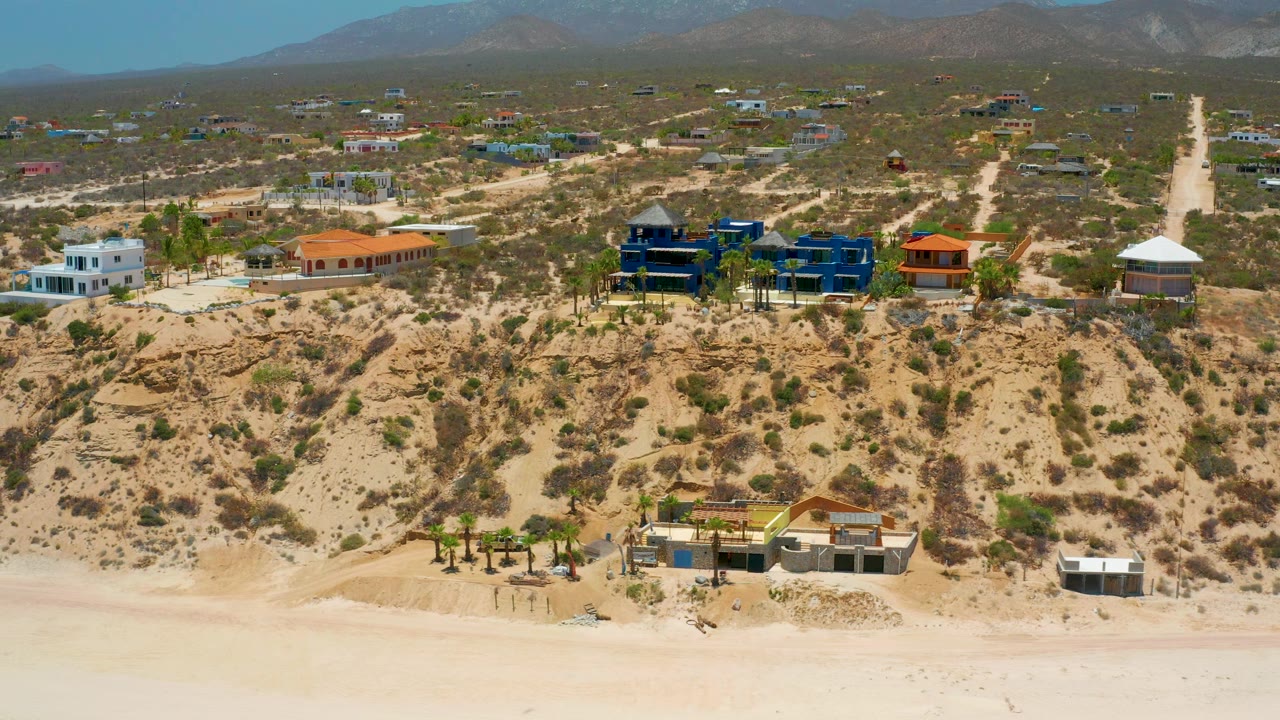 Aerial frame over El Sargento La Ventana, Mexico, with the beach band across the bottom, scrub-covered slope, low modern homes, a straight dirt road climbing inland, mountains, and no visible people.
