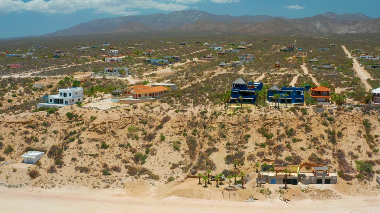 Opening aerial frame of El Sargento La Ventana, Mexico, with pale beach edge, scrubby hillside homes, dirt roads, distant mountains, blue sky, and no visible people.