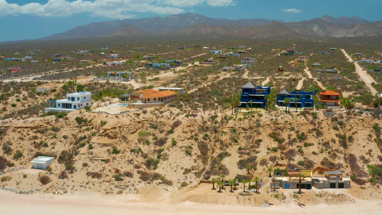 Midwide aerial frame in El Sargento La Ventana, Mexico, with blue homes near center right, a white house to the left, pale dirt tracks, mountains behind, and no visible people.