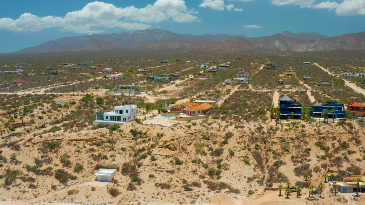 Aerial view over El Sargento La Ventana, Mexico, showing sandy scrubland, scattered homes, dirt roads, distant mountains, bright clouds, and no visible people.