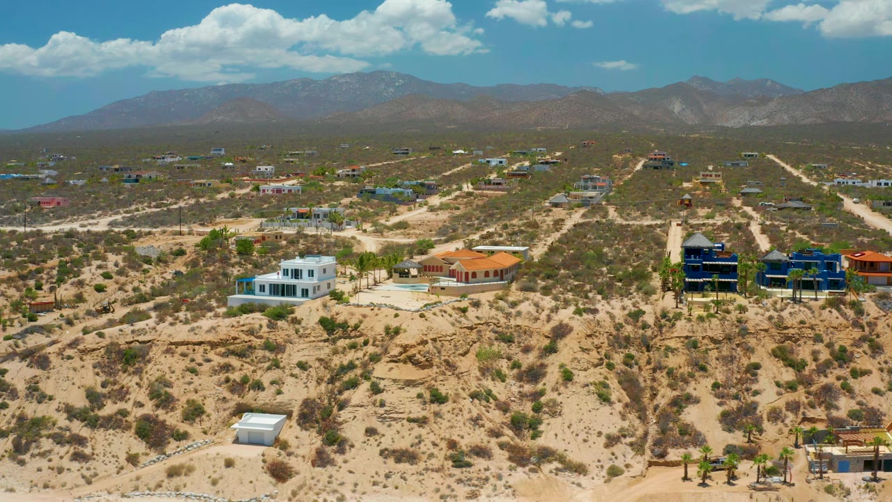 Drone-height view across El Sargento La Ventana, Mexico, with a white house, orange-roofed building, blue buildings, unpaved roads, mountain backdrop, and no visible people.