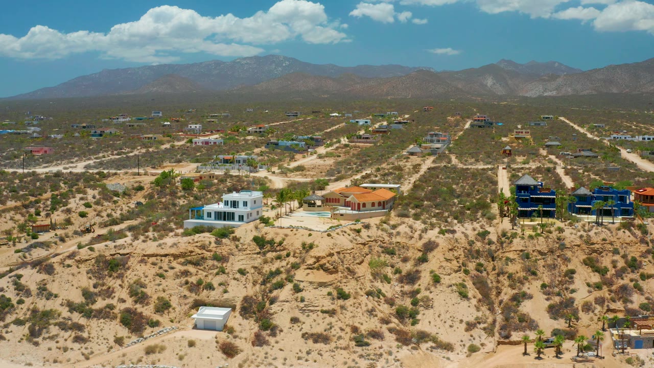 Wide aerial frame in El Sargento La Ventana, Mexico, showing dry tan terrain, spaced houses, straight dirt roads, mountains under clouds, and no visible people.