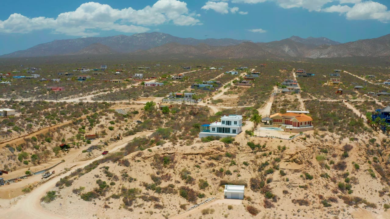 Aerial frame over El Sargento La Ventana, Mexico, showing the white foreground house, nearby orange-roofed building, desert road grid, mountains, and no visible people.