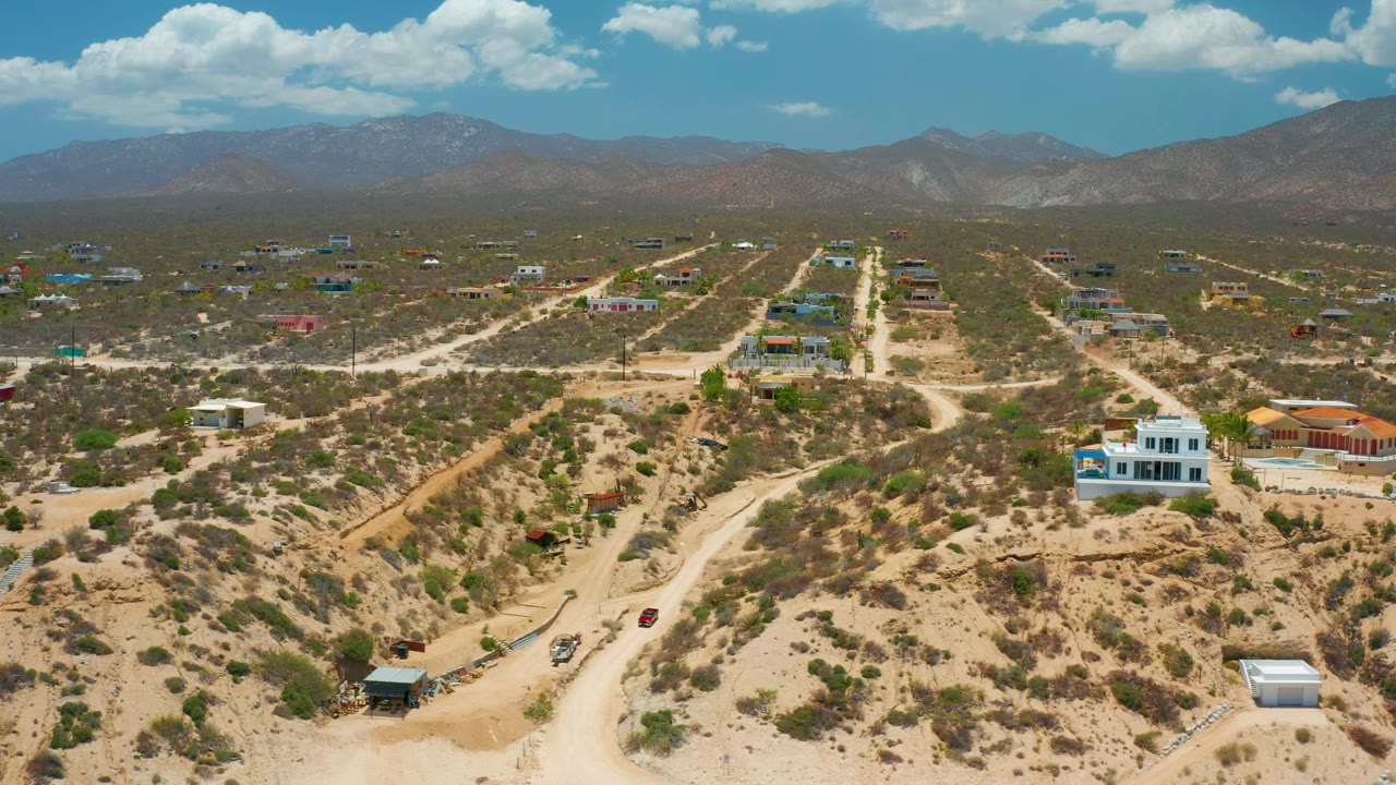 Drone-height view of El Sargento La Ventana, Mexico, with a white house near the lower right, sandy tracks through scrub, distant homes, mountain horizon, and no visible people.