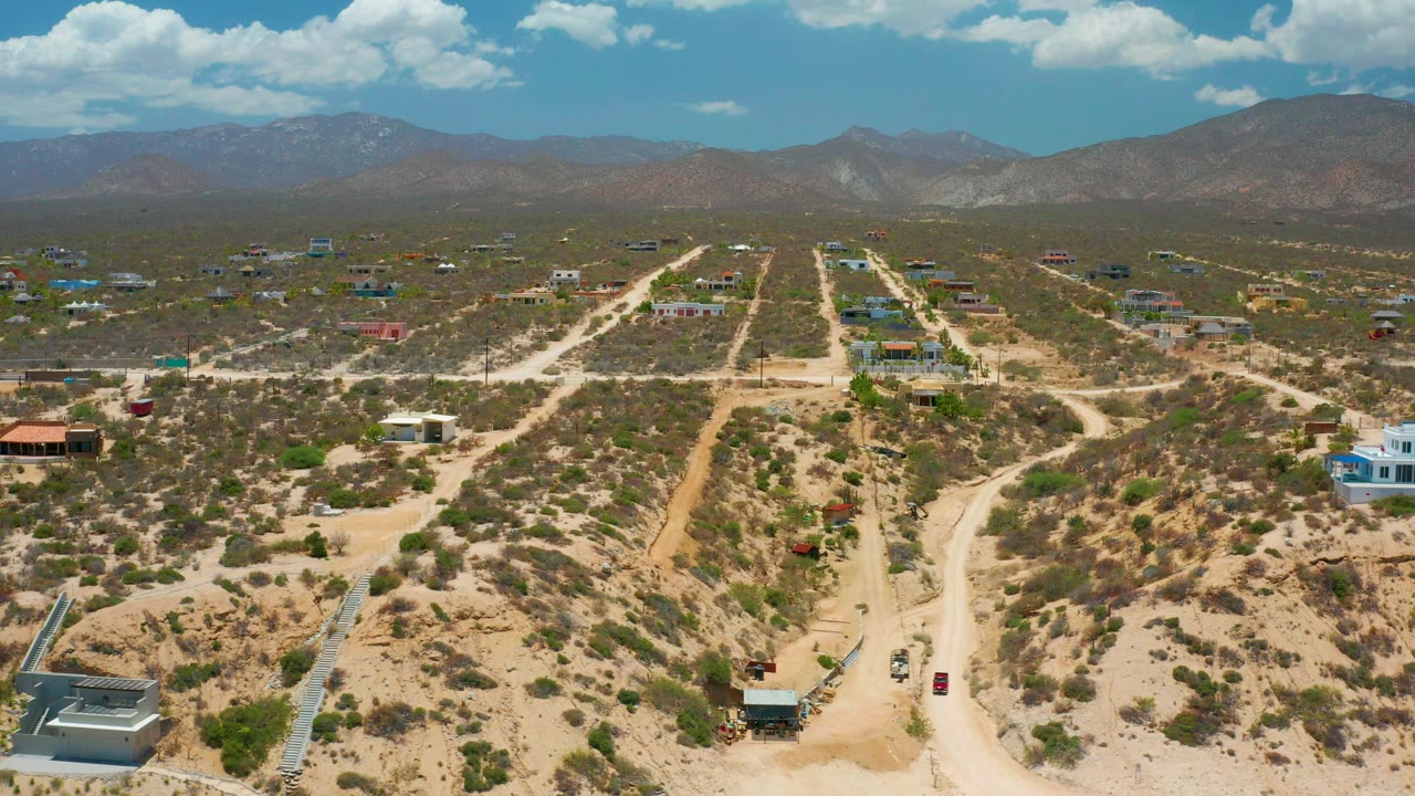 Opening frame of El Sargento La Ventana, Mexico, showing sandy roads through scrubland, scattered buildings, distant mountains, a blue sky with white clouds, and no visible people.