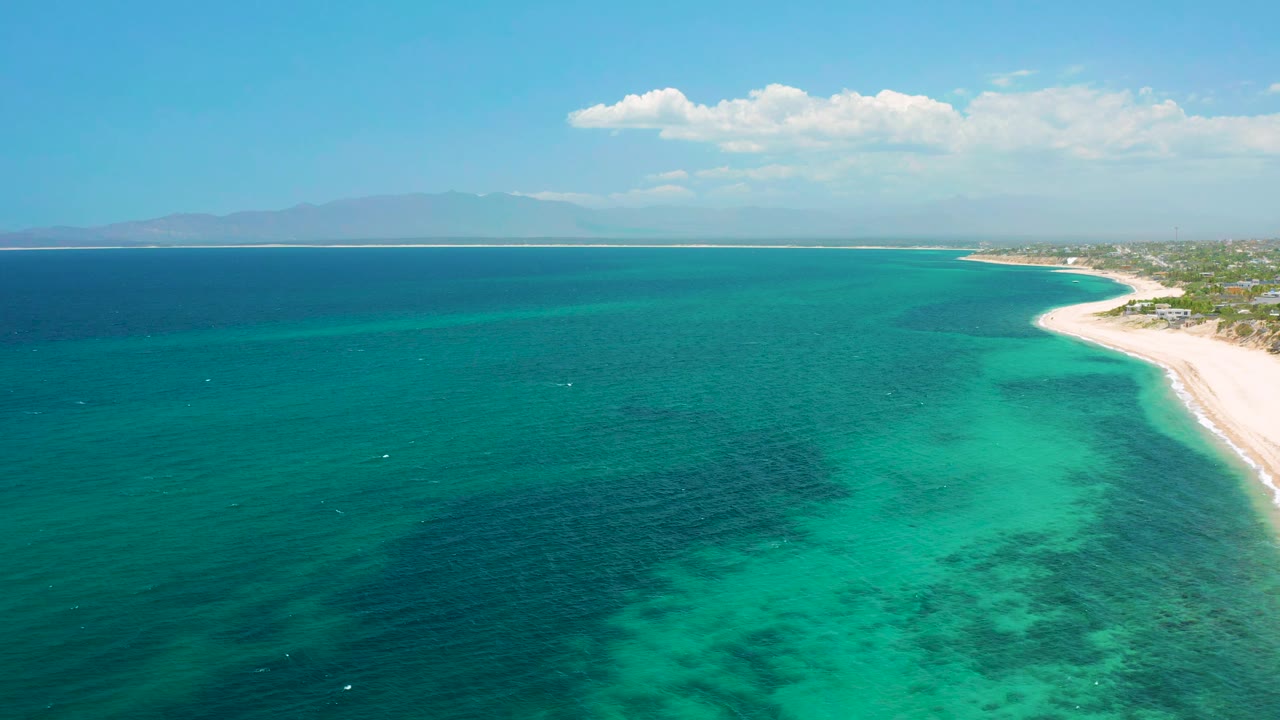 Aerial frame at El Sargento La Ventana, Mexico, showing darker blue water to the left, bright shallow turquoise patches below, the beach along the right edge, and no visible people.