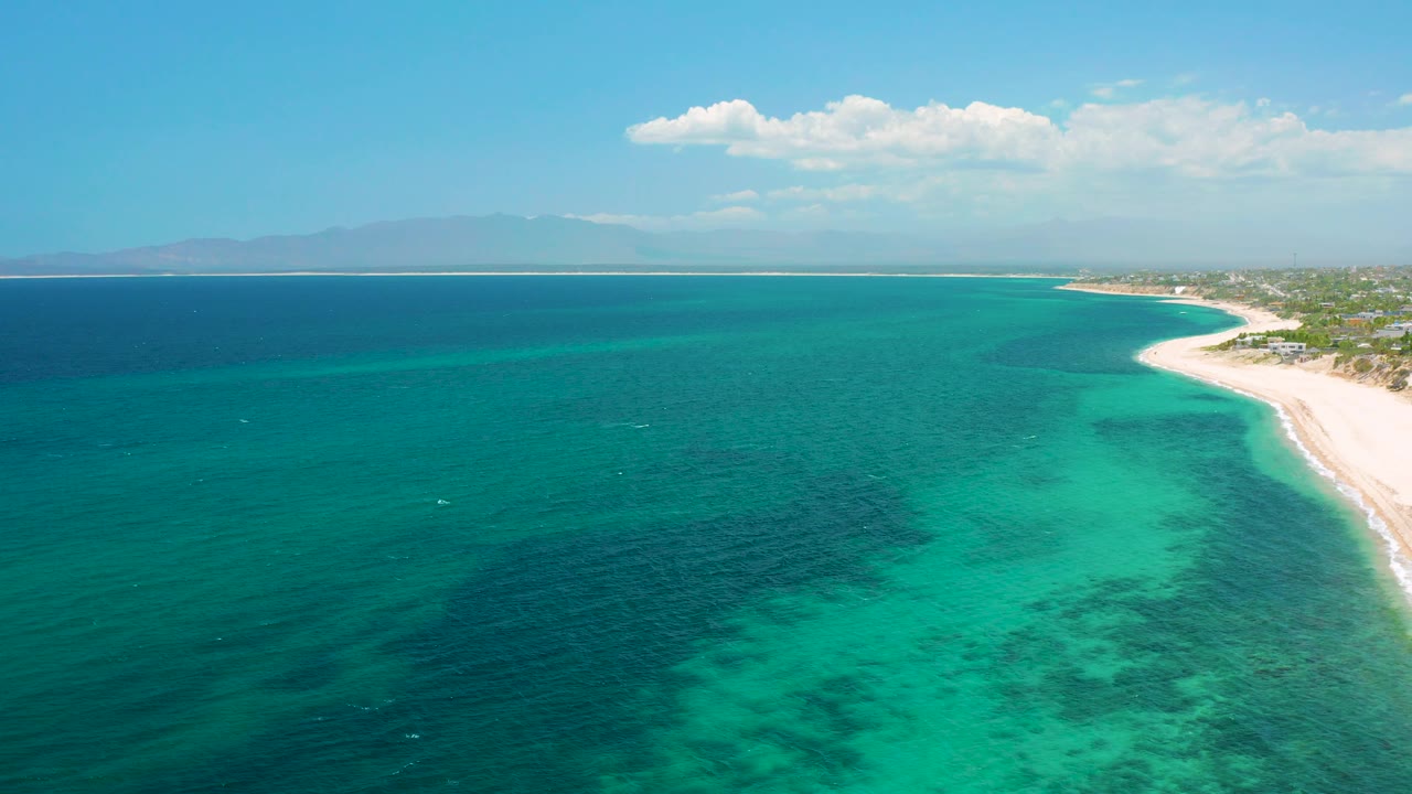 Wide aerial coastline with calm sea, pale sand, and distant land.