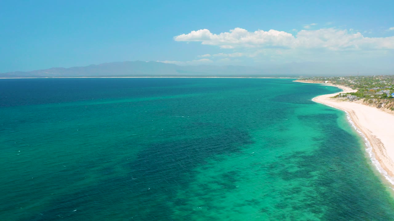 Aerial frame from El Sargento La Ventana, Mexico showing turquoise water in the foreground, a pale beach curving along the right side, distant mountains, white clouds, and no visible people.