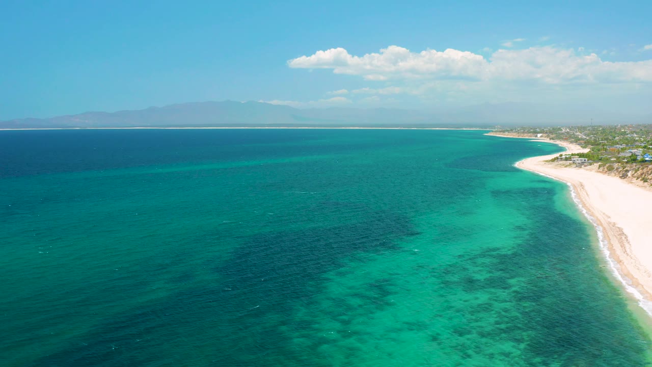 Aerial view in El Sargento La Ventana, Mexico showing a wide sweep of teal water, a bright sand strip on the right, small coastal buildings beyond it, hazy mountains, and no visible people.