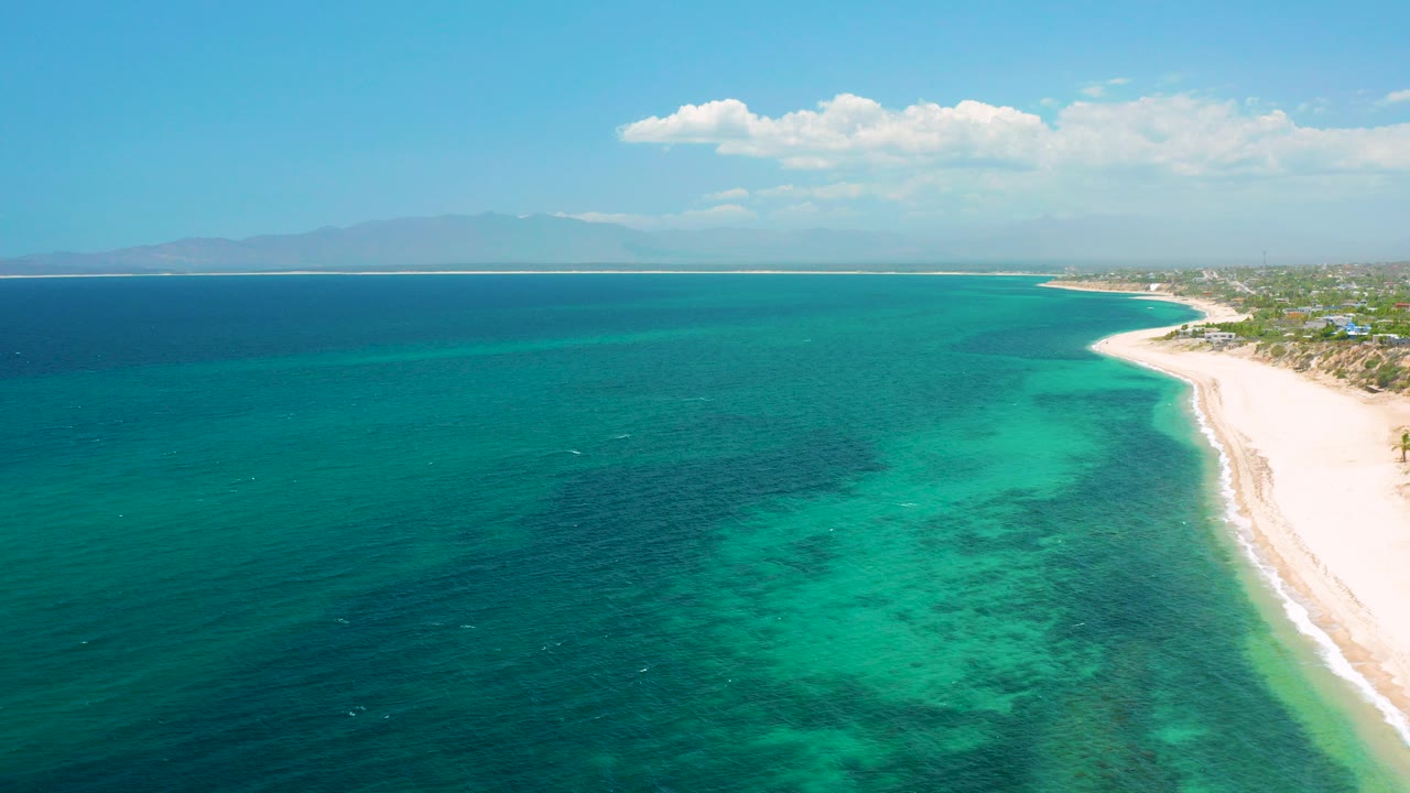 Aerial view over El Sargento La Ventana, Mexico, with deep blue water at left, turquoise shallows near a pale beach at right, distant mountains, and no visible people.