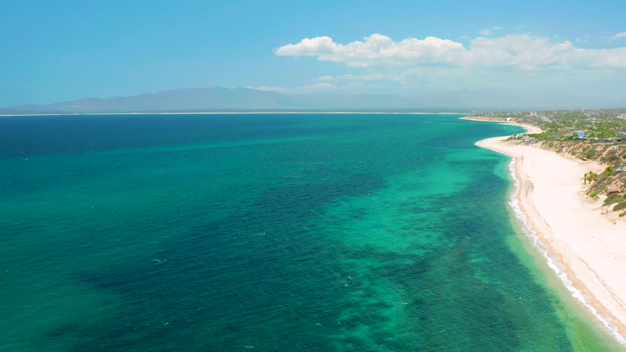 Aerial view over El Sargento La Ventana, Mexico, showing turquoise ocean beside a pale curving beach, low clouds, distant mountains, and no visible people.