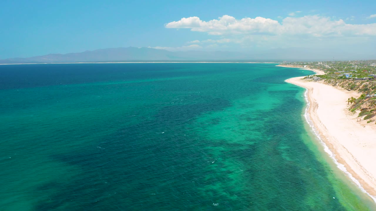 Aerial coastline at El Sargento La Ventana, Mexico, with shallow turquoise water, deeper blue water offshore, a curving white beach, scattered clouds, and no visible people.